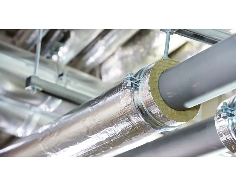 A close-up image of industrial pipes wrapped with foil-backed insulation material, likely Rockwool mineral wool, secured with metal clamps. The insulation covers the pipes, leaving a section exposed to show the inner material. The setting appears to be an